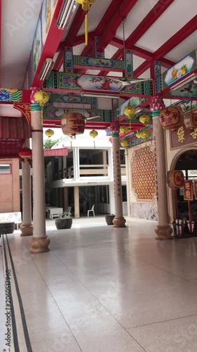The interior of an ornately decorated Chinese Buddhist temple in Thailand. The ceiling is decorated with bright mosaics, lanterns and hanging plants. 