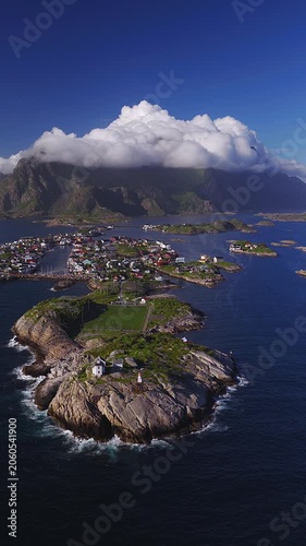 Aerial view of Henningsvaer football pitch in Lofoten, Norway, with lighthouse, skerries, docks, boats, village streets, and jagged mountains under rolling clouds.