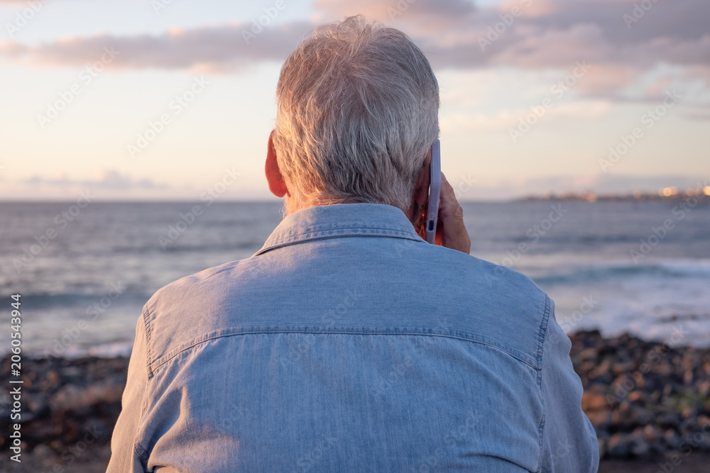 Fototapeta premium Senior man with gray hair standing on the beach at sunset, holding a smartphone to his ear and looking toward the ocean, relaxed and connected during retirement