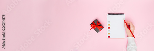 Female hand writing on blank notepad with gift box and red ribbon on pastel pink background, festive flat lay banner with copy space for holiday promotion, planning, goals, to-do list or wish list