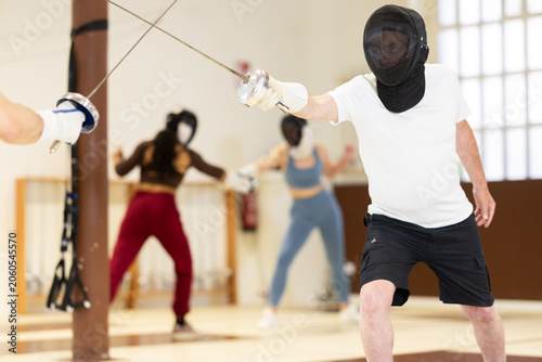 Tableau sur toile Elderly man in a protective helmet holds a rapier and participates in a training