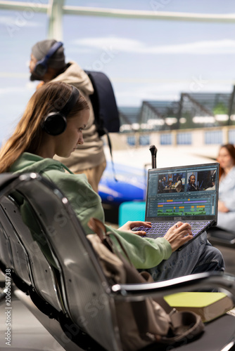 Fotografie Digital nomad filmmaker cutting video clips and applying color grading on laptop in the airport terminal, preparing web content during travel, Remote creative work and global mobility on a trip