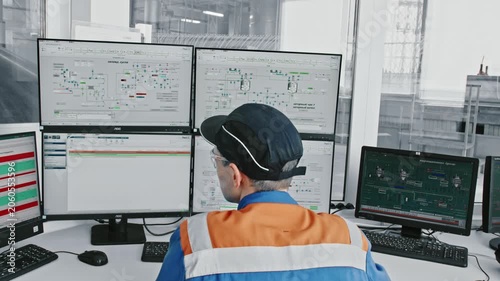 Industrial engineer in a control room, sitting at a desk with multiple monitors, supervising automated manufacturing processes and checking technical data on computer screens for quality control