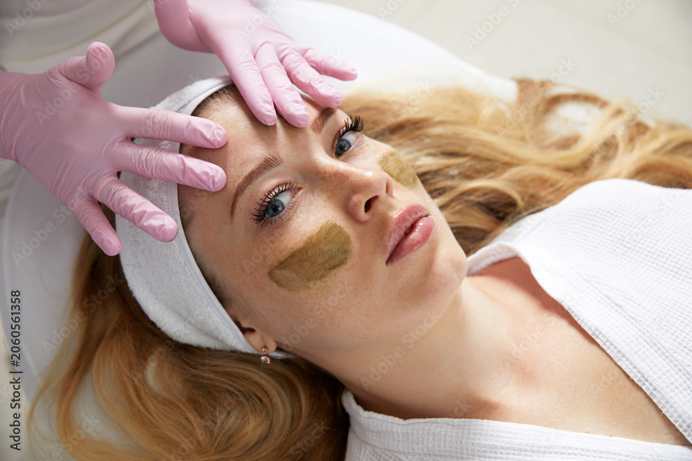Naklejka premium Cosmetologist applying a mask from clay to the face of a beautiful woman. Spa treatment and face care in the beauty salon