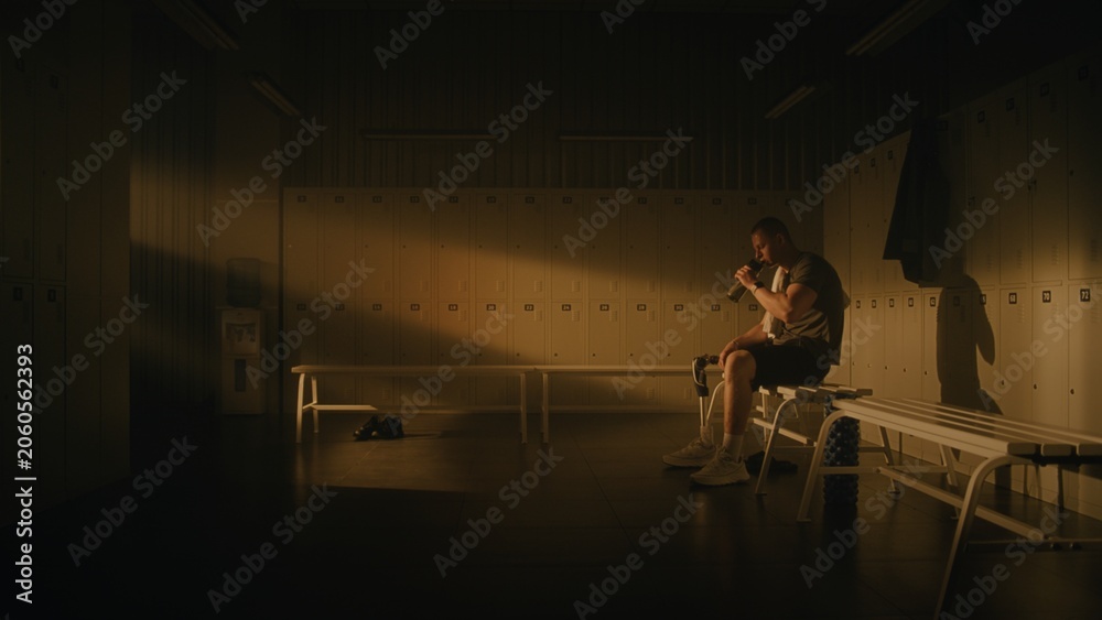 Fototapeta premium Reality of Hard Work. Tired Young Man With Prosthetic Leg Sits on Locker Room Bench After Intense Workout. Male Takes Sip of Water and Wipes Face With Towel, Resting in Warm Glow of Gym Setting Sun.