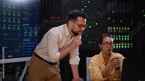 It specialists analyzing data on a laptop and large screen while working together in a modern server room at night