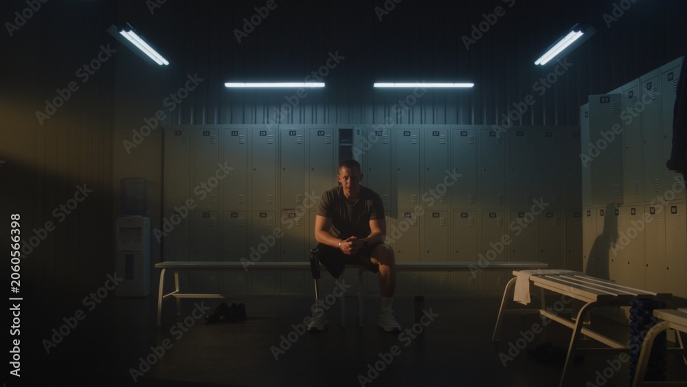 Fototapeta premium Young Man with Prosthetic Leg Sits on Bench in Dimly Lit Locker Room, Focusing Mind and Building Motivation Before Standing Up to Start Workout Session. Mental Grit and Determination. Portrait.