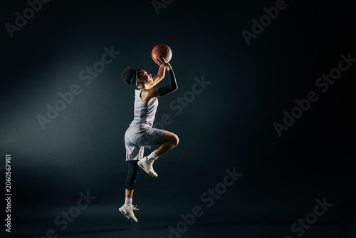 A female basketball player in team uniform jumps with a ball in a studio