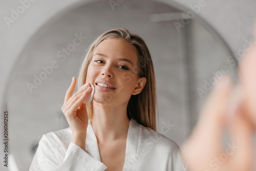Fotografie Smiling Young Woman With Light Skin Applying Skincare Cream To Her Cheek In Front Of Bathroom Mirror