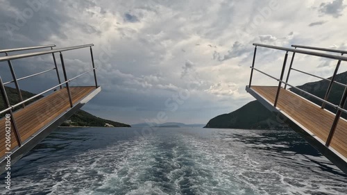 Rear view from a passenger ferry cruising through the Ionian Sea near Lefkada, Greece. White wake stretches across deep blue water between open stern platforms, with mountains and dramatic clouds 
