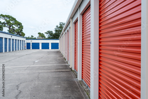 A storage outside facility with multiple units featuring red roll-up doors on one side and blue doors on the other, aligned in neat rows along a paved driveway