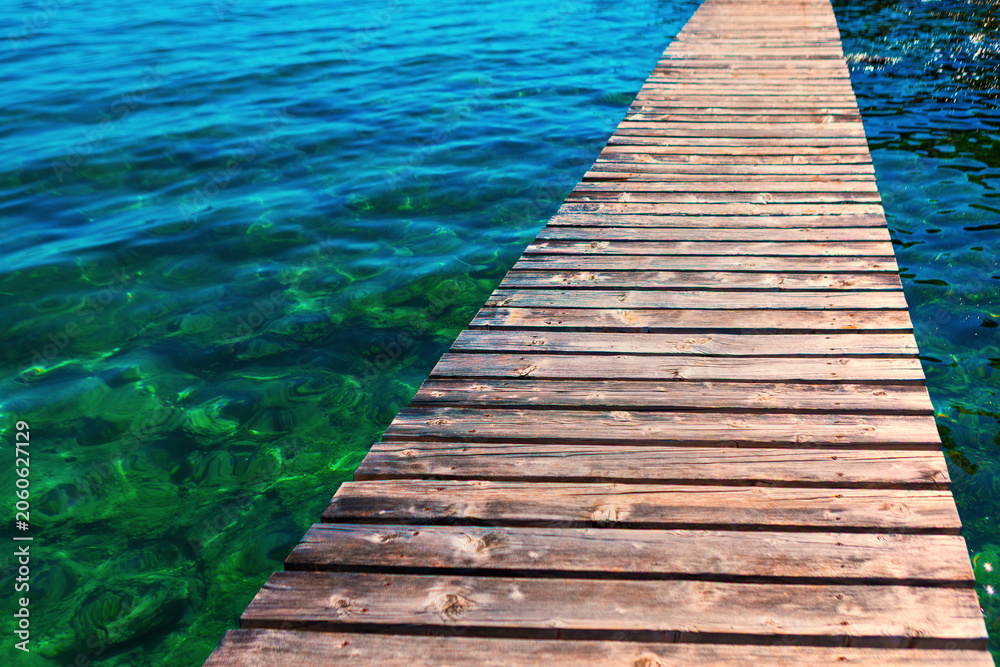 Fototapeta premium Weathered wooden dock extends diagonally over strikingly clear water, blending vivid turquoise and emerald green hues. Sunlight filters through the shallow depths, revealing rocky lakebed below