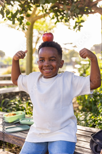African American child sitting on bench in park balancing apple, flexing arms, lunchbox, backpack