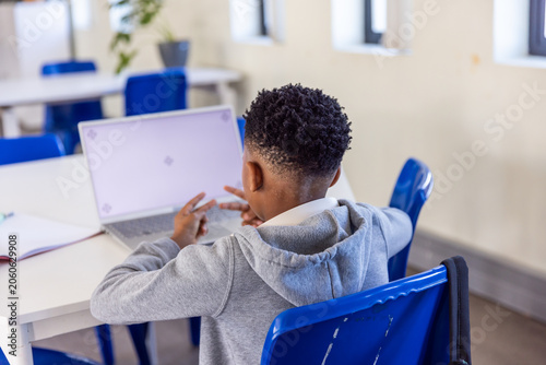 African American child wearing gray hoodie sitting at classroom table using laptop and notebook