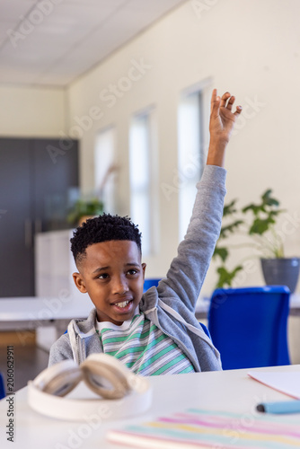 African American schoolboy raising hand while sitting at desk in class with headphones and notebook