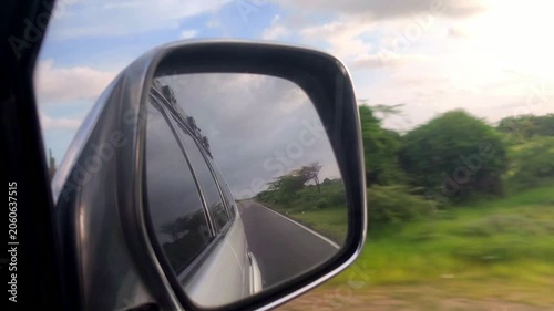 Close-up of a car side mirror reflecting the vehicle and road while driving past blurred green countryside, conveying travel, motion, and speed on a scenic route.