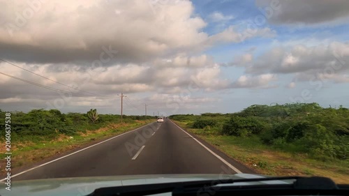 Wide view from a moving car along a quiet paved highway through green countryside, with a distant vehicle, roadside vegetation, and dramatic cloudy sky.