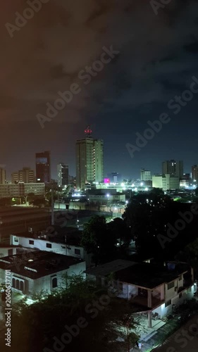 Urban skyline at night with glowing office towers, apartment buildings, and street lights under a dark cloudy sky. A moody cityscape showing downtown lights and residential rooftops.
