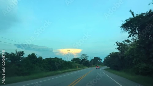 Empty rural highway curving through lush green trees under a blue evening sky, with a glowing sunset cloud on the horizon and a vehicle in the distance.