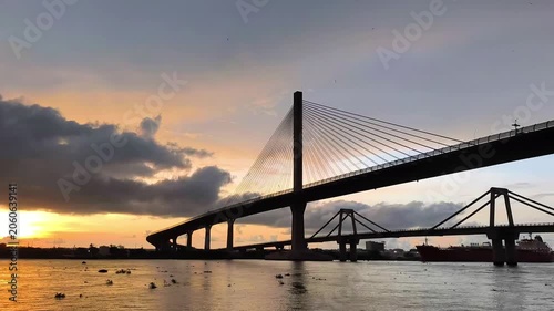 Dramatic sunset view of a cable-stayed Pumarejo bridge silhouetted against glowing clouds above calm water, with reflections and city skyline details creating a serene urban landscape.