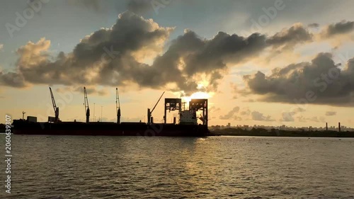 Silhouette of cranes and port infrastructure against a dramatic sunset sky, with golden light reflecting on calm water. A moody industrial seascape with clouds and distant city skyline.