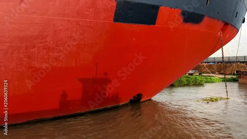 Close-up of a red ship hull with weathered texture, visible waterline and reflections on the surface, creating an abstract maritime background with industrial details.