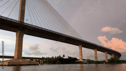 Wide view of a large cable-stayed bridge crossing a calm river at sunset, with dramatic lines, tropical shoreline, and soft evening sky in the background.
