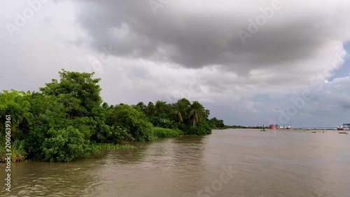 Lush tropical vegetation lines a calm riverbank beneath dramatic storm clouds, creating a moody natural landscape with water, greenery, and overcast weather.