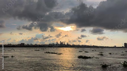 Moody sunset over a distant city skyline reflected on calm water, with dark clouds and warm golden light creating a dramatic urban landscape scene.