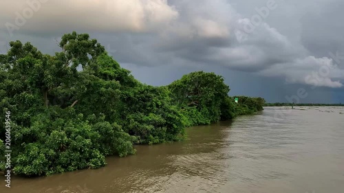 Dense tropical trees line a muddy riverbank beneath heavy storm clouds, creating a dramatic natural landscape with calm water and lush green vegetation.