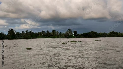 Wide river under dramatic storm clouds with a tropical tree-lined shoreline, calm flowing water, and a moody natural atmosphere before rain.