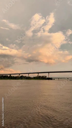 Dramatic golden clouds glow above a wide river with a long bridge on the horizon, capturing a calm sunset landscape with warm light and reflective water.