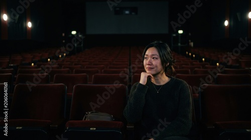 Emotional young Asian woman crying alone in a dark, empty movie theater with rows of red seats and a large screen