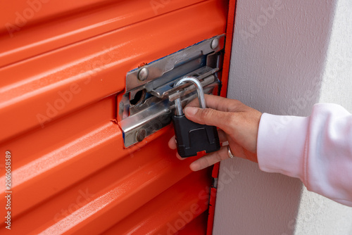 A hand in a white sleeve unlocks a black padlock on a metal latch attached to an orange corrugated storage door, indicating entry to a secured storage unit