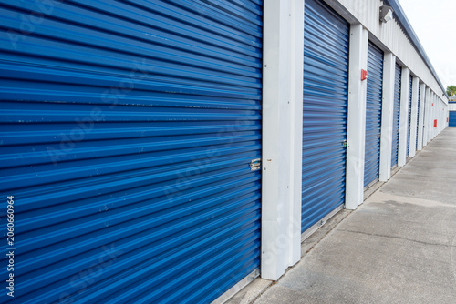 A row of storage units with blue corrugated metal doors and white frames, aligned along a paved corridor that leads into the distance, emphasizing an organized and structured facility