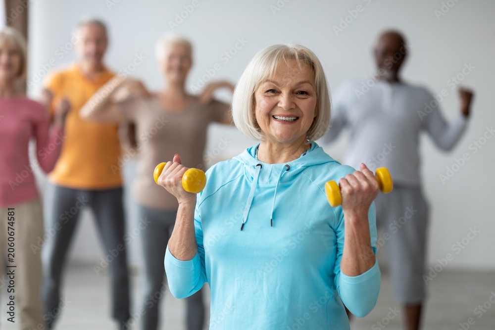Naklejka premium Smiling Senior Caucasian Woman Participating In Group Exercise Class, Lifting Dumbbells With Confidence And Joy, Engaged In Fitness Activities In Bright Modern Studio. Weight Loss Concept.