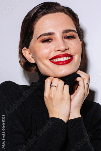 Confident brunette woman poses for a fashion beauty portrait with a warm smile, bold red lipstick and black turtleneck, holding the collar near her face against a bright studio background.