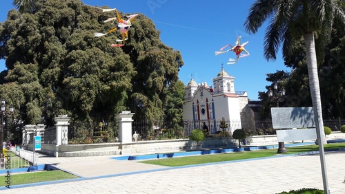 Принти на полотні Town square in Oaxaca Mexico featuring a white colonial church, the famous Tule tree, colorful star-shaped piñatas hanging in the air, and a blue-trimmed fountain under a clear blue sky