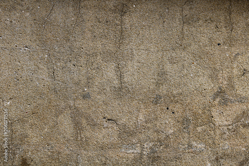 An old, weathered concrete wall surface. The rough, granular facade is covered with fine vertical and horizontal cracks, and subtle dark stains from moisture and age.