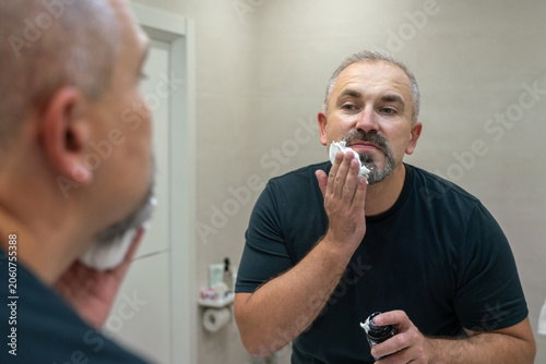 Portrait of handsome gray-haired middle-aged man applying shaving foam to trim his beard