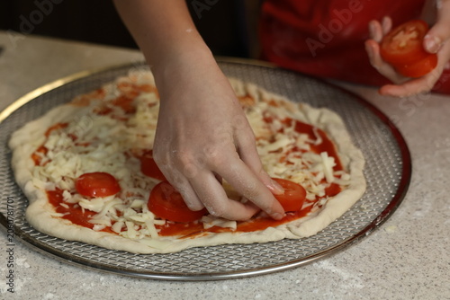 Tablou pe pânză The process of making homemade pizza: a woman's hands arrange slices of fresh tomatoes and peppers onto a pizza base that has already been sprinkled with cheese