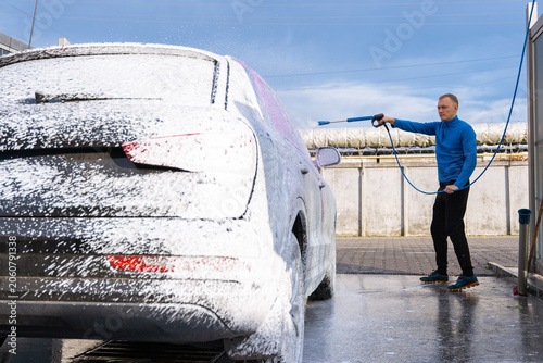 Man Washing Car at Self Service Car Wash. Applying thick foam with a pressure washer. The scene takes place in an outdoor washing bay with modern self-service equipment, automotive accessories and