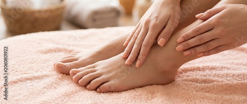 Female hands with manicured nails gently touching bare feet on a soft pink towel, with a blurred background of spa elements and towels in a serene indoor setting