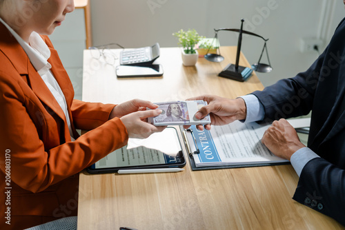 Fotografie Close view of a cash payment exchanged across a lawyer’s desk with legal papers, gavel and scales in the background, suggesting bribery, corruption or an illegal business deal