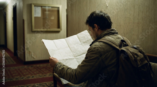 Lost backpacker studying paper map in dim hotel hallway with worn walls luggage nearby budget travel uncertainty navigation check in challenge concept confusing accommodation low cost lodging