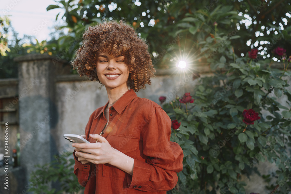 Obraz premium Joyful woman enjoys an outdoor lifestyle moment, smiling while using a smartphone in a sunny garden, wearing a rust red blouse among flowers, capturing candid warmth and charm