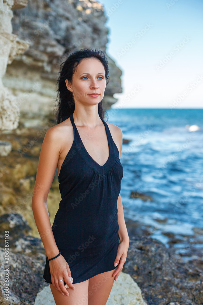 Fototapeta premium Slim Brunette Woman in Black Dress on Rocky Sea Coast in Summer