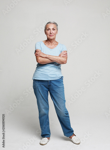 Mature woman standing over grey background. Old senior female, 60s gray-haired lady looking at camera arms crossed.
