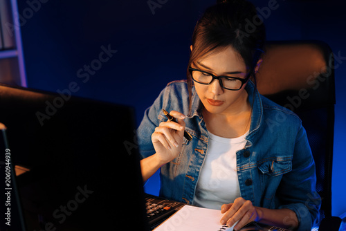 Tablou pe pânză Thinking young beautiful Asian creative woman looking on pc with taking note on paper for memo in sales target report on business market graph report at neon light modern office at night