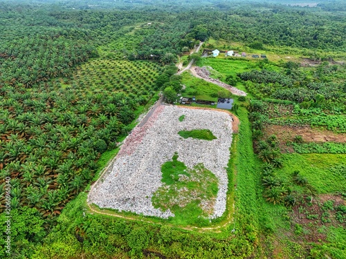 Aerial view of a large waste dump site surrounded by lush greenery and palm tree plantations.